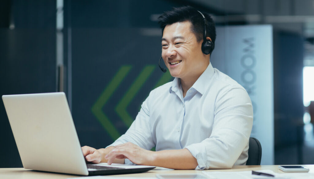 Portrait of a smiling young Asian man working in the office at a laptop in a headset